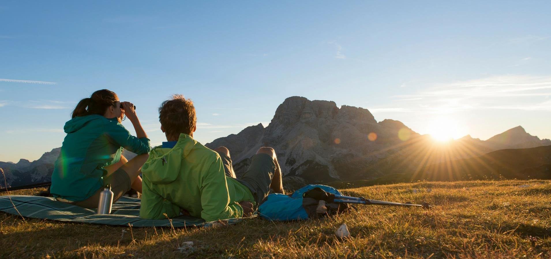 Due escursionisti osservano il tramonto sulle montagne seduti su una coperta.