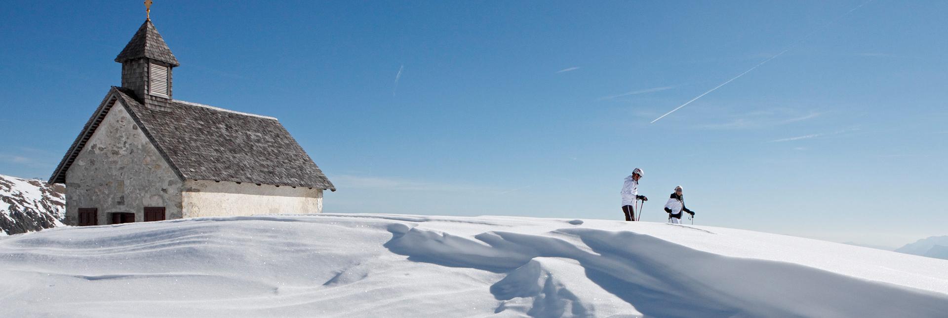 Due scialpinisti presso una cappella in un paesaggio invernale innevato sotto cielo blu.